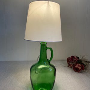 Może przedstawiać: Zielona szklana lampa z białym abażurem. Podstawa lampy to butelka z uchwytem. Klosz ma jasnokremowy kolor i lekko rozszerzający się kształt. Lampa jest podłączona i świeci.