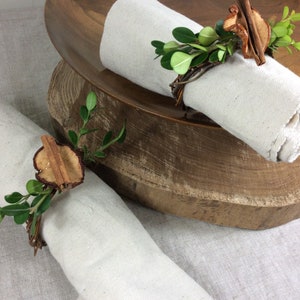 Może przedstawiać: Dwa białe lniane serwetki z obrączkami do serwetek z cynamonu i suszonych plastrów jabłka. Obrączki są ozdobione zielonymi gałązkami.