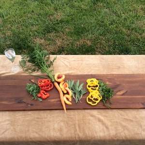 May include: A long, rectangular wooden serving board with a handle on one end. The board is stained a dark brown color and has a natural wood grain. The board is topped with sliced vegetables, including red bell peppers, carrots, peaches, and yellow bell peppers. There are also sprigs of fresh herbs on the board.