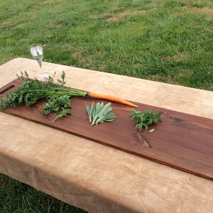 May include: A long, rectangular wooden serving board with a brown stain. The board has a handle on one end and is decorated with fresh herbs and a carrot.