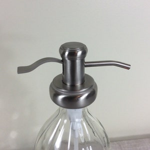 May include: A stainless steel soap dispenser pump with a clear glass bottle. The pump has a lever and a spout.