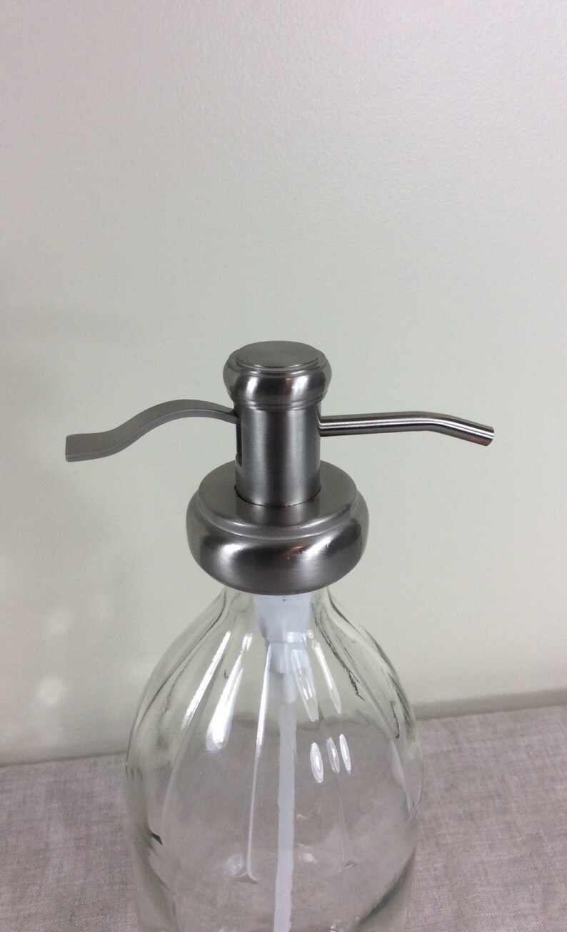 May include: A stainless steel soap dispenser pump with a clear glass bottle. The pump has a lever and a spout.