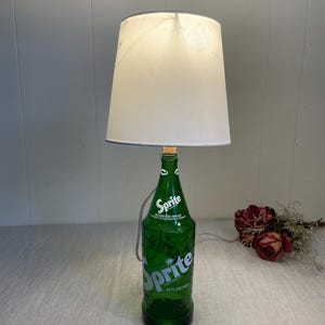 May include: A green Sprite bottle lamp with a white lampshade. The bottle features the Sprite logo in white and the text "32 fl oz (1qt)". The lampshade is white with a blue trim.