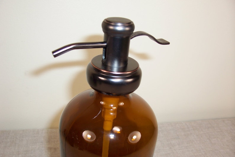 May include: A brown glass bottle with a black and copper pump dispenser. The bottle has a textured surface and is designed for dispensing liquids.