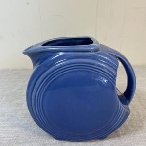 May include: A blue ceramic pitcher with a unique, rounded shape and a handle. The pitcher has a spout and a square opening at the top. The body of the pitcher features concentric, ridged detailing. The pitcher is a solid, vibrant blue color.