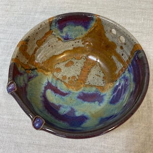 May include: A handmade ceramic bowl with a unique, organic design. The bowl features a blend of colors, including brown, purple, and teal, with a speckled, textured surface. It has a small spout and two small handles.