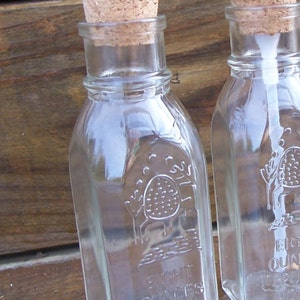 May include: Two clear glass bottles with cork stoppers. The bottle on the left has a label that reads "Eight Ounces Pure Honey".