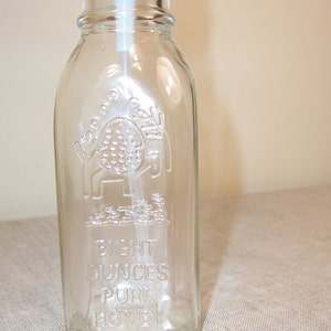 K&ouml;nnte beinhalten: Eine durchsichtige Glasflasche mit einem Korkstopfen und einem Pumpspender. Die Flasche ist mit den Worten "Eight Ounces Pure Honey" gepr&auml;gt.