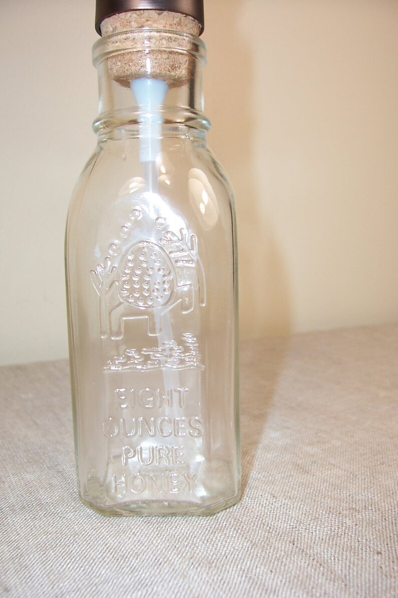K&ouml;nnte beinhalten: Eine durchsichtige Glasflasche mit einem Korkstopfen und einem Pumpspender. Die Flasche ist mit den Worten "Eight Ounces Pure Honey" gepr&auml;gt.