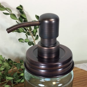 May include: A bronze soap dispenser pump with a clear glass jar. The pump has a long, curved spout and a round, smooth top.