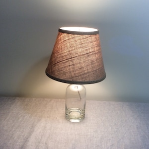 May include: A table lamp with a brown burlap lampshade and a clear glass bottle base.