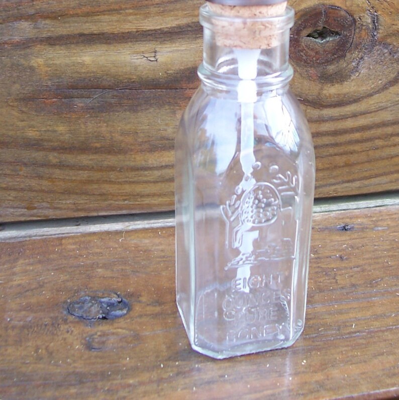 K&ouml;nnte beinhalten: Eine klare Glasflasche mit einem Korkverschluss. Die Flasche ist mit den Worten "Eight Ounces Pure Honey" gepr&auml;gt.