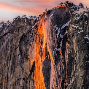 May include: A fiery orange waterfall cascades down a rocky cliff face, illuminated by the setting sun. The cliff is covered in snow and ice, creating a dramatic contrast with the warm colors of the waterfall.