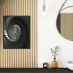 May include: A black and white photograph of a spiral staircase in a modern bathroom setting. The photo is framed and hanging on a wall with wood paneling. A round mirror with a wooden frame hangs on the wall above a white porcelain sink.