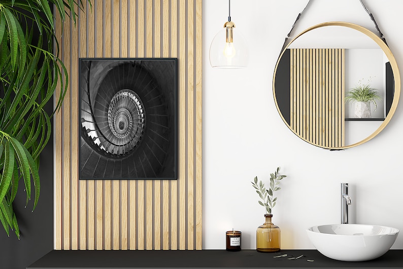 May include: A black and white photograph of a spiral staircase in a modern bathroom setting. The photo is framed and hanging on a wall with wood paneling. A round mirror with a wooden frame hangs on the wall above a white porcelain sink.