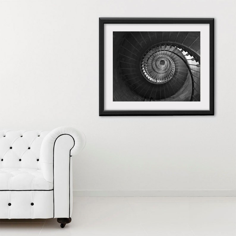 May include: Black and white photograph of a spiral staircase framed in a black frame. The photo is hanging on a white wall.