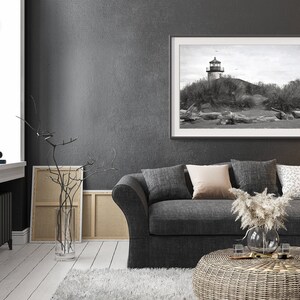 May include: A black and white photograph of a lighthouse on a sandy beach. The photograph is framed in a black frame and hanging on a gray wall. A gray couch with pillows and a wicker coffee table are in the foreground.