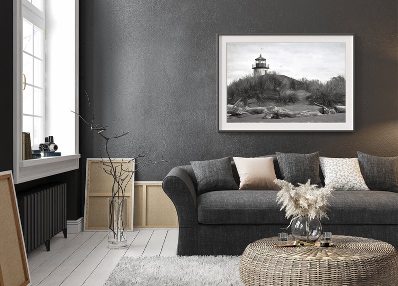 May include: A black and white photograph of a lighthouse on a sandy beach. The photograph is framed in a black frame and hanging on a gray wall. A gray couch with pillows and a wicker coffee table are in the foreground.