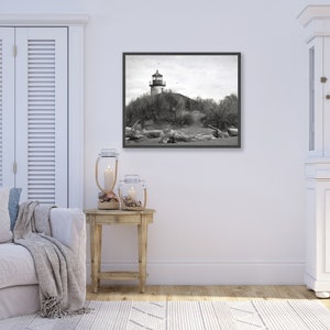 May include: A black and white photograph of a lighthouse on a sandy beach. The lighthouse is tall and white with a black roof. The beach is covered in sand and driftwood. The photo is framed in a black frame.