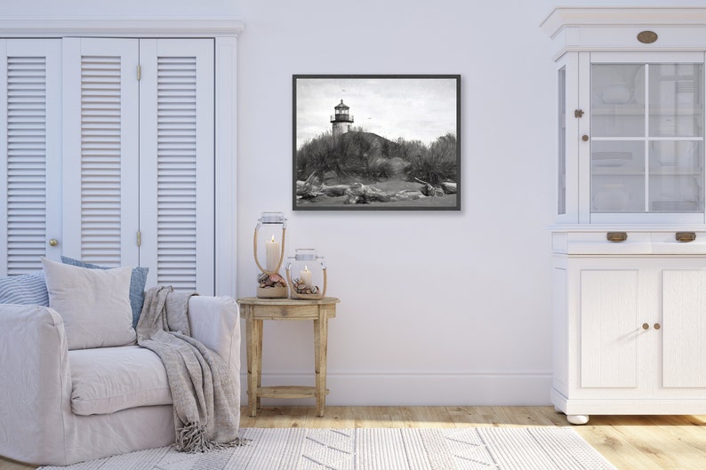 May include: A black and white photograph of a lighthouse on a sandy beach. The lighthouse is tall and white with a black roof. The beach is covered in sand and driftwood. The photo is framed in a black frame.