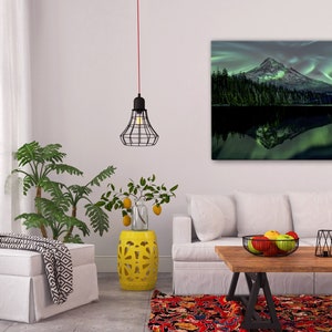 May include: A living room interior design with a white couch, a yellow ceramic stool, a white ottoman, a wooden coffee table, a black metal wire pendant light, and a framed canvas print of a mountain with a green lake and a green aurora borealis reflection.