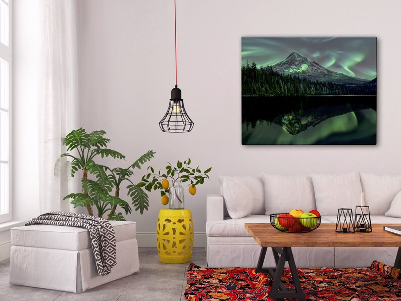 May include: A living room interior design with a white couch, a yellow ceramic stool, a white ottoman, a wooden coffee table, a black metal wire pendant light, and a framed canvas print of a mountain with a green lake and a green aurora borealis reflection.