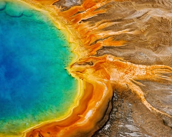 May include: Aerial view of a vibrant hot spring with a bright blue center surrounded by a yellow and orange rim. The surrounding landscape is a mix of brown and orange.
