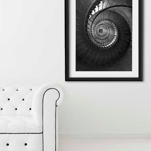 May include: Black and white photograph of a spiral staircase framed in a black frame. The photo is hanging on a white wall above a white sofa.