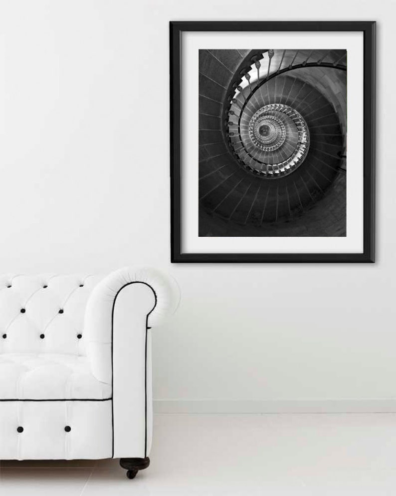 May include: Black and white photograph of a spiral staircase framed in a black frame. The photo is hanging on a white wall above a white sofa.