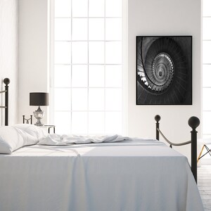 May include: A white bed with a black metal frame and a white headboard. The bed is made with white sheets and a white blanket. There is a black and white photo of a spiral staircase hanging on the wall behind the bed. There is a white chair in the corner of the room.