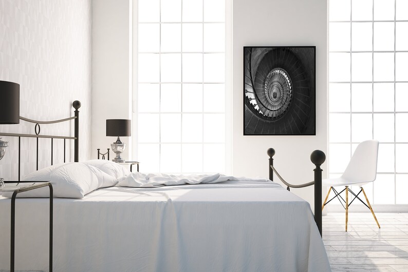May include: A white bed with a black metal frame and a white headboard. The bed is made with white sheets and a white blanket. There is a black and white photo of a spiral staircase hanging on the wall behind the bed. There is a white chair in the corner of the room.