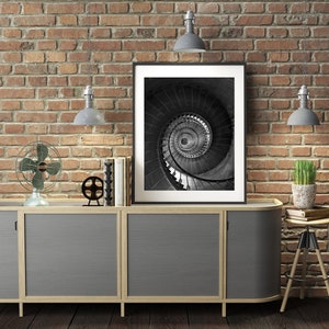 May include: A gray cabinet with three doors and a wood frame sits in front of a brick wall. A black and white photo of a spiral staircase hangs above the cabinet. There are two lamps on the wall, a fan on the cabinet, and a plant on a stool to the right of the cabinet.