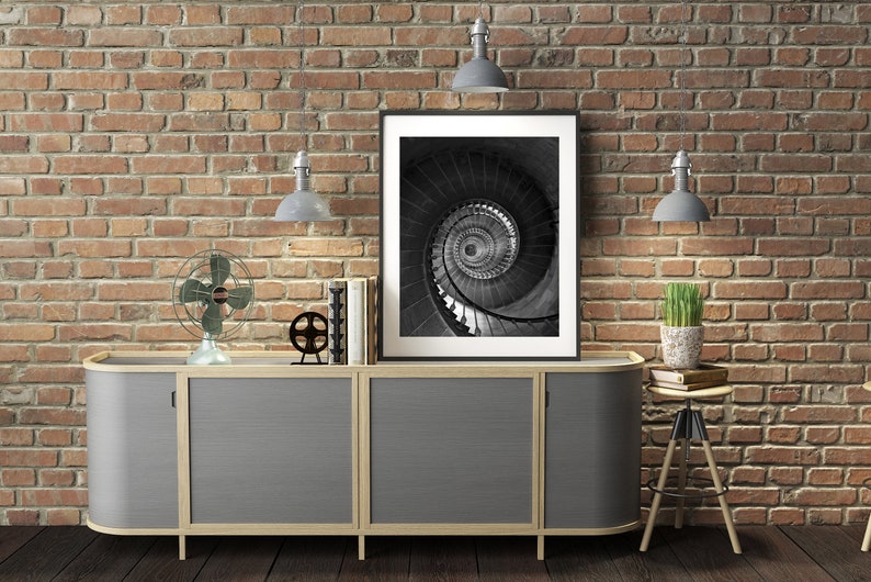 May include: A gray cabinet with three doors and a wood frame sits in front of a brick wall. A black and white photo of a spiral staircase hangs above the cabinet. There are two lamps on the wall, a fan on the cabinet, and a plant on a stool to the right of the cabinet.