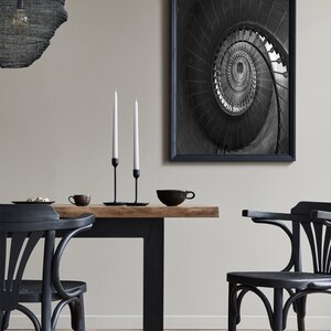 May include: A black and white photograph of a spiral staircase. The photo is framed in a black frame and is hanging on a wall above a black dining table with two black chairs. There are two white candlesticks on the table.