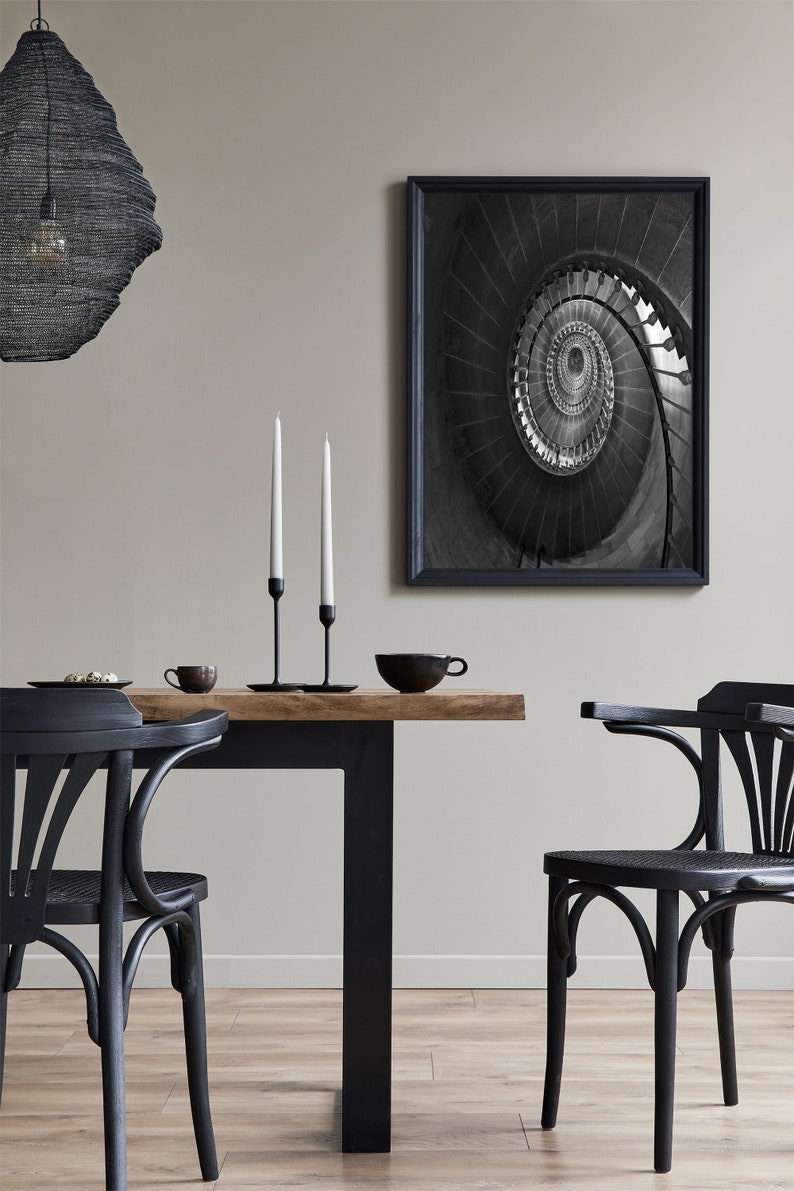 May include: A black and white photograph of a spiral staircase. The photo is framed in a black frame and is hanging on a wall above a black dining table with two black chairs. There are two white candlesticks on the table.
