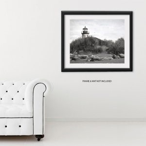 May include: Black and white photograph of a lighthouse on a sandy beach with driftwood in the foreground. The lighthouse is tall and white with a black roof and a black and white striped tower. The beach is sandy and the water is calm.