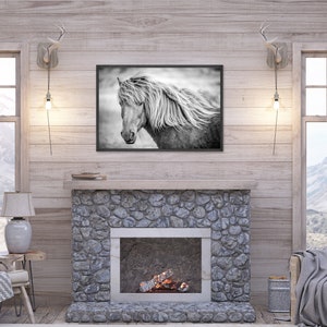 May include: A black and white photograph of a horse's head with its mane blowing in the wind, framed and hanging on a wall above a stone fireplace in a rustic living room.