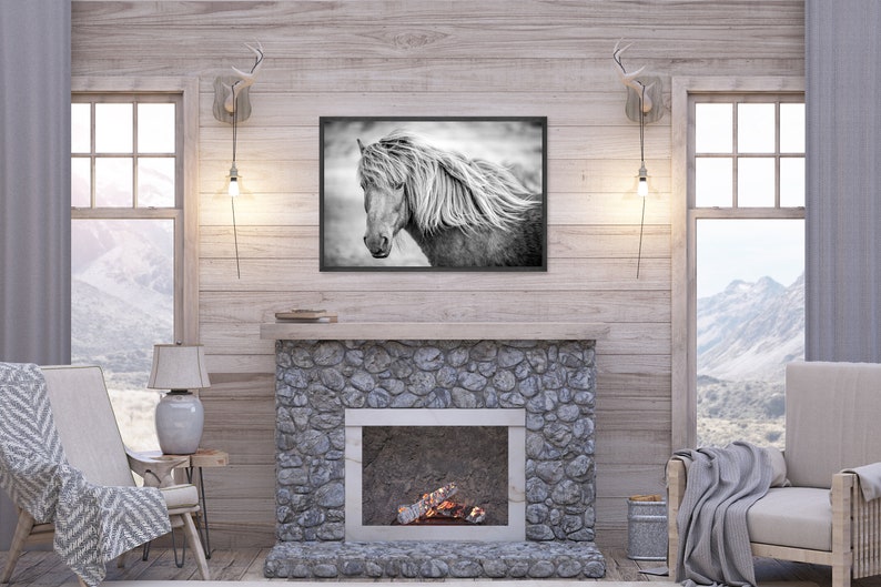 May include: A black and white photograph of a horse's head with its mane blowing in the wind, framed and hanging on a wall above a stone fireplace in a rustic living room.