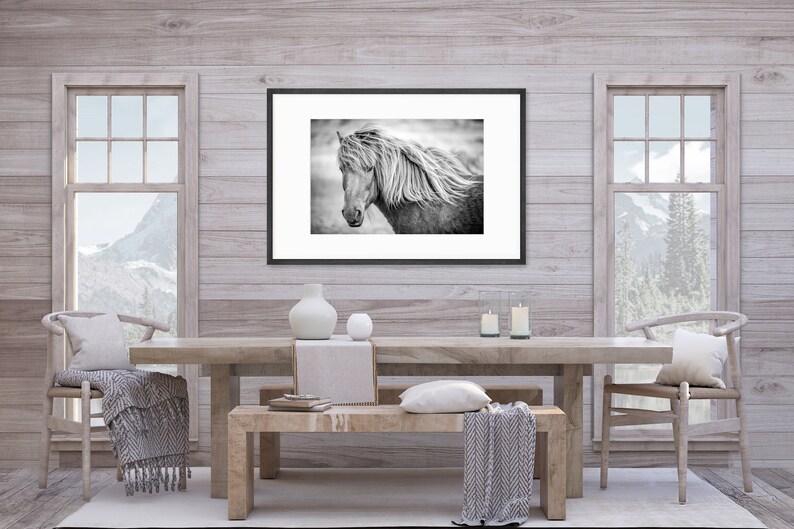 May include: A black and white photograph of a horse with a long flowing mane, framed in a black frame and hanging on a light wood wall. The photo is above a wooden table with two chairs and a bench.