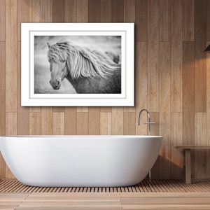 May include: Black and white photograph of a horse with a long flowing mane, framed in a white frame and hanging on a wood wall above a white bathtub.