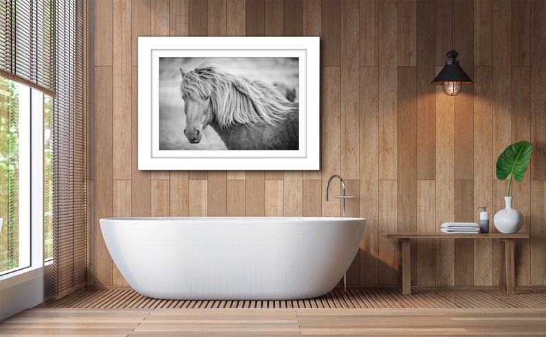 May include: Black and white photograph of a horse with a long flowing mane, framed in a white frame and hanging on a wood wall above a white bathtub.