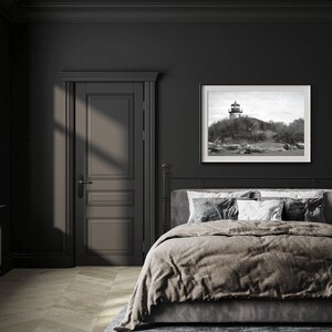 May include: A black and white photograph of a lighthouse on a rocky shore hangs above a bed with a brown duvet cover and white pillows in a dark room with a closed door.