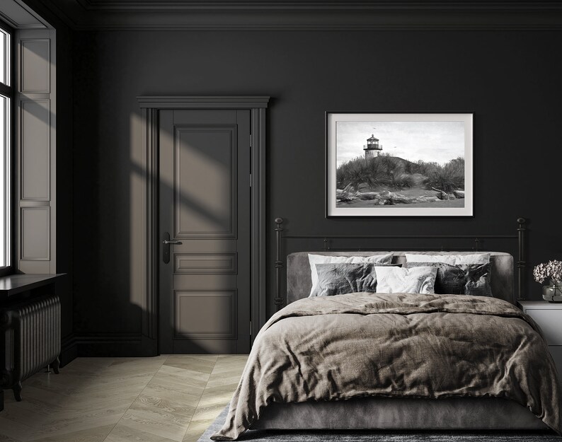 May include: A black and white photograph of a lighthouse on a rocky shore hangs above a bed with a brown duvet cover and white pillows in a dark room with a closed door.