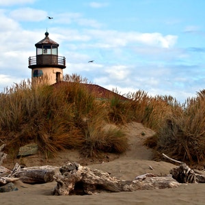 Pode incluir: Um farol desgastado ergue-se numa praia de areia, rodeado de ervas altas. O farol &eacute; pintado de branco com um telhado castanho e tem uma pequena janela no lado. O c&eacute;u &eacute; azul com nuvens brancas.