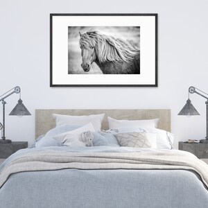 May include: A black and white photograph of a horse's head with a flowing mane. The horse is looking to the left of the frame. The photo is framed in a black frame and hanging on a white wall above a bed.