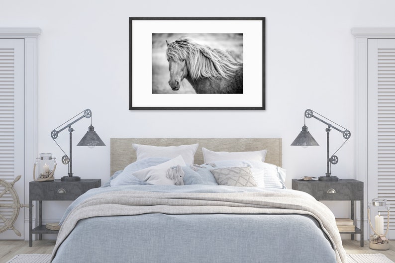 May include: A black and white photograph of a horse's head with a flowing mane. The horse is looking to the left of the frame. The photo is framed in a black frame and hanging on a white wall above a bed.