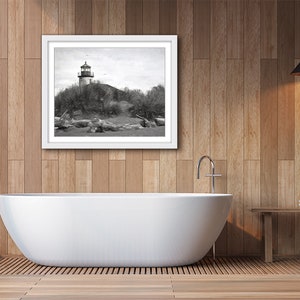 May include: A black and white photograph of a lighthouse on a rocky shore. The photo is framed in a white frame and hanging on a wood-paneled wall above a white bathtub.