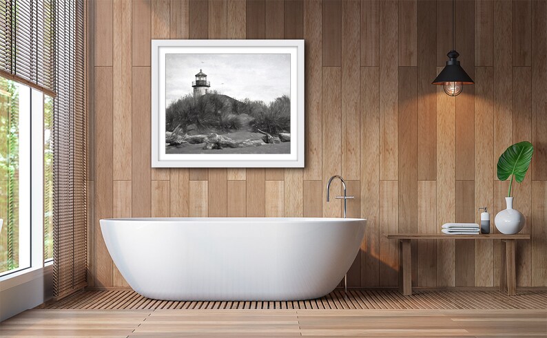 May include: A black and white photograph of a lighthouse on a rocky shore. The photo is framed in a white frame and hanging on a wood-paneled wall above a white bathtub.