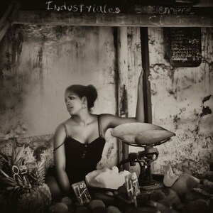 May include: A woman in a black tank top sits in front of a vintage scale with a wooden frame. The scale has a metal pan and a dial with numbers. The woman is looking to the side and has her arm resting on the scale. The background is a brick wall with the words "Industriales" and "Siempre" written on it.