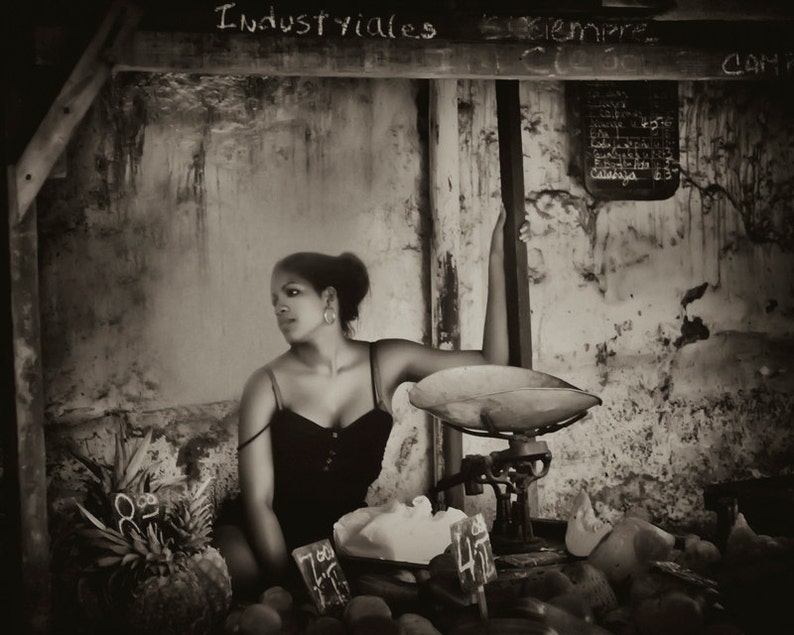 May include: A woman in a black tank top sits in front of a vintage scale with a wooden frame. The scale has a metal pan and a dial with numbers. The woman is looking to the side and has her arm resting on the scale. The background is a brick wall with the words "Industriales" and "Siempre" written on it.